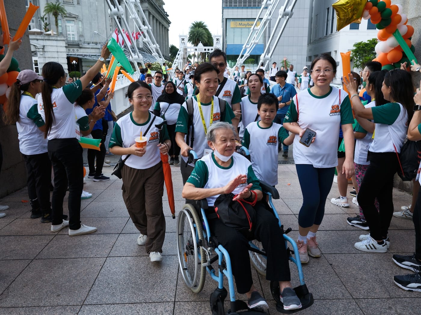 Crowd wearing "SPD Walkathon" shirts, cheering elderly woman in wheelchair on a city street. Maybank logo visible.